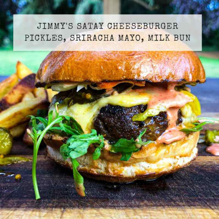 A picture of the recipe for Jimmy's Satay Cheeseburger with pickles and sriracha mayo and a toasted bun with chips in the background.