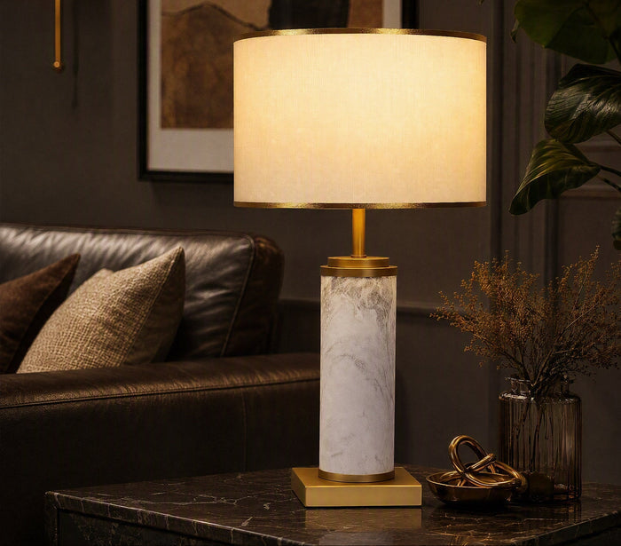 Illuminated white marble cylinder table lamp on dark marble side table beside leather sofa