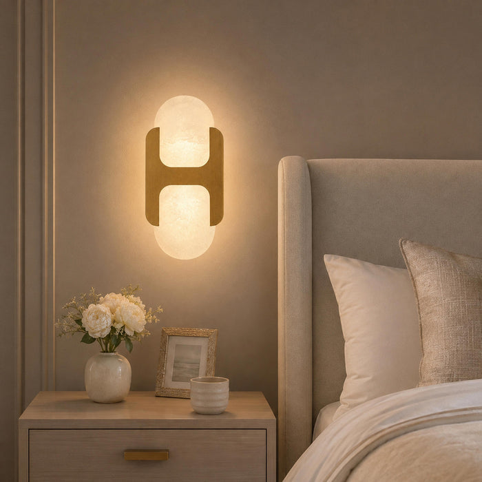 Warm illuminated H-shaped alabaster wall sconce mounted beside a upholstered bedhead above a nightstand in a cozy bedroom